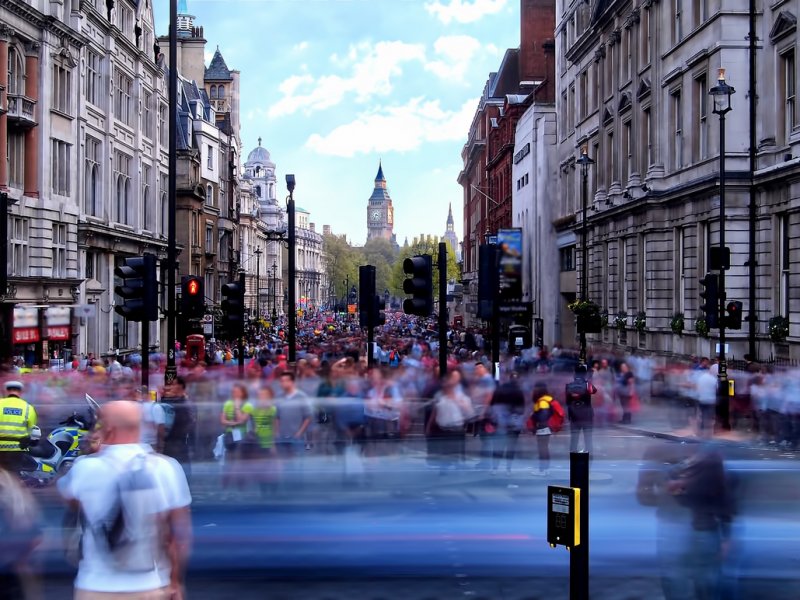 Long exposure in the afternoon of a crowd of blurred people walking along Whitehall towards Trafalgar Square as a police man stands by a motorbike (left).
